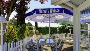 Een buitenzitplaats met een tafel en stoelen onder een blauwe parasol met de tekst 'Maisel's Weisse'.