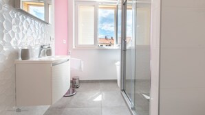 A modern bathroom with a white vanity, a glass shower enclosure, and a view of buildings through a window. - Apartman Tonci (Primosten)