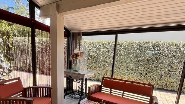 Un área de estar exterior moderna con mobiliario de madera y un gran ventanal con vistas a un seto.