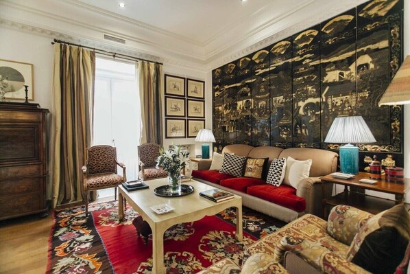 Een woonkamer met een rode bank, gestreept fauteuils, een bijzettafeltje met een vaas, en een muur met ingelijste kunstwerken.