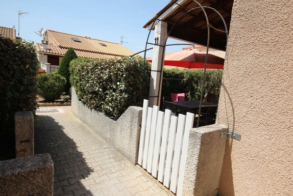 A paved pathway leading to a building with a white fence and a red umbrella. - Corner villa located in a secured residence with pool, Airco, new renovated, 400m from the beach... (PORTIRAGNES)