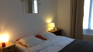 A bedroom with a bed, a nightstand with a lamp, a dresser, and a window with curtains. - Apartment-cottage of the church (Châteauneuf-sur-Loire)