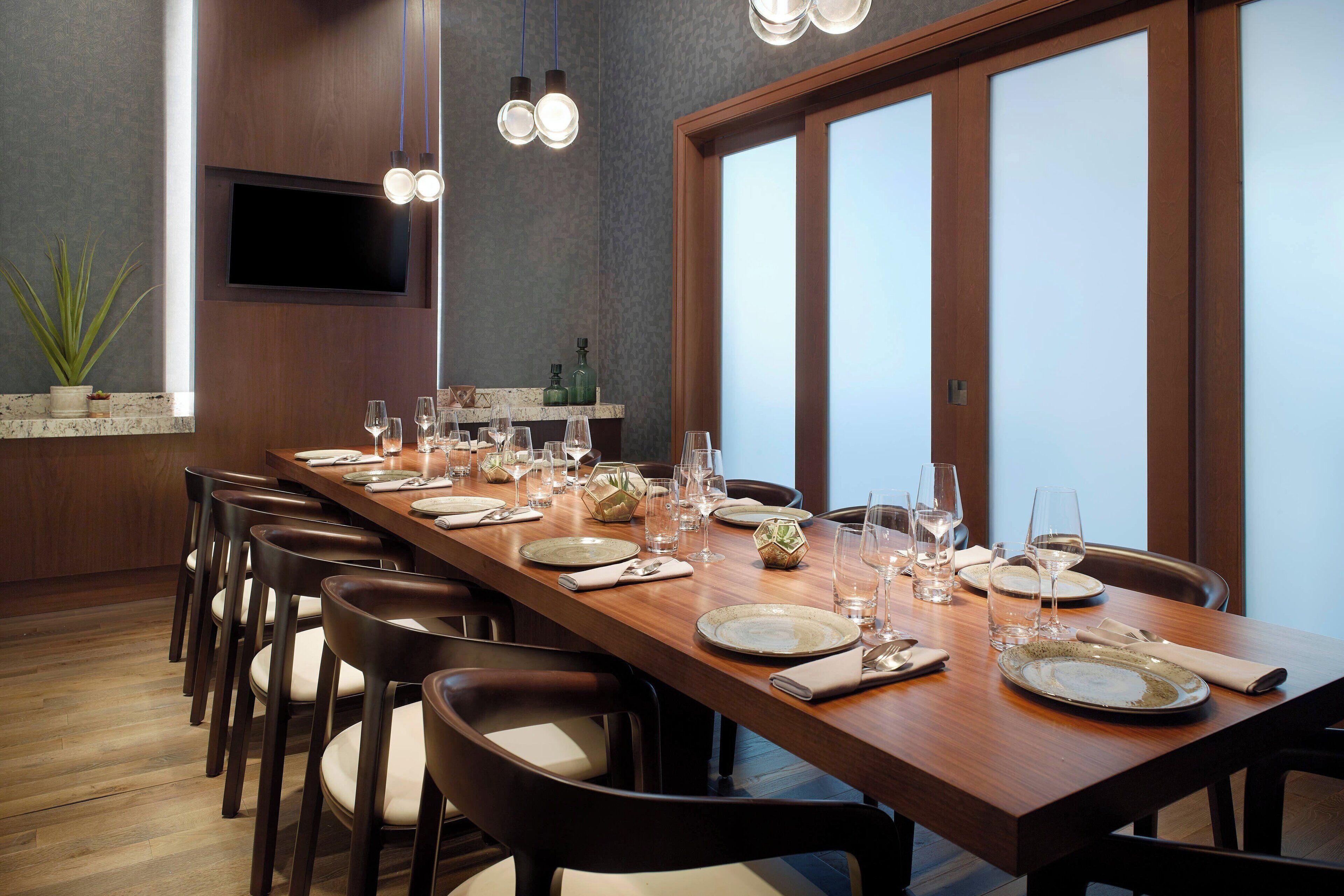 A long dining table set with plates, glasses, and chairs in a modern room with a TV and pendant lights.