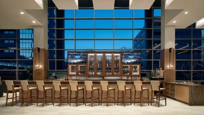 Un bar moderno con una barra alargada, diversas opciones de asientos y grandes ventanales con vistas al centro de la ciudad.