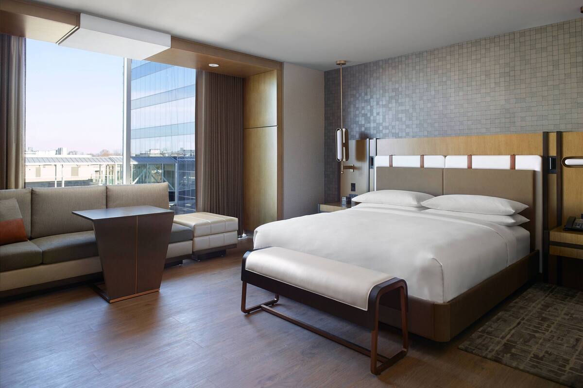 A modern hotel room with a large bed, a sofa, a small table, and a view of the city through large windows.