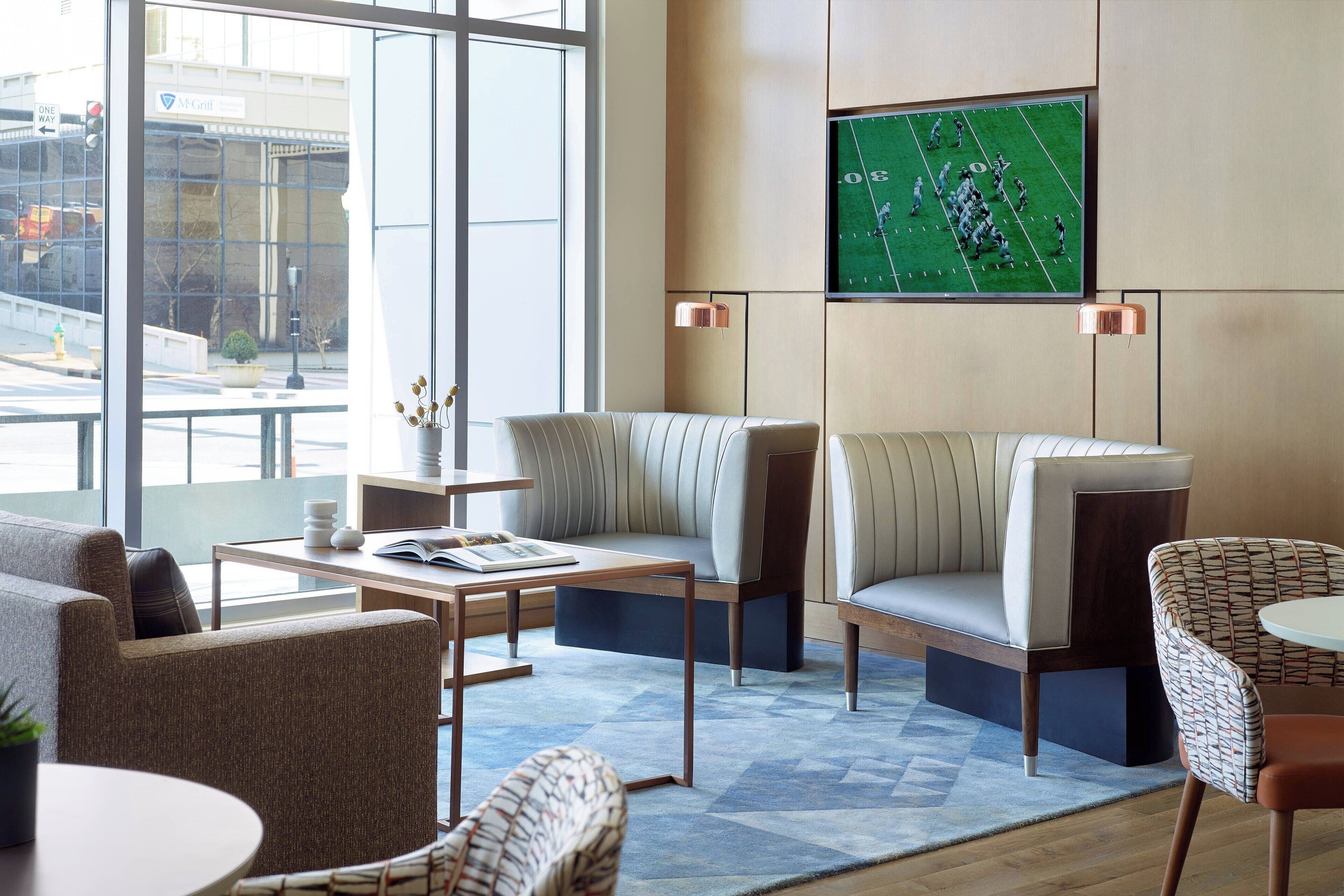 A modern hotel lobby with seating arrangements, a flat-screen TV mounted on the wall, and a view of the city through large windows.