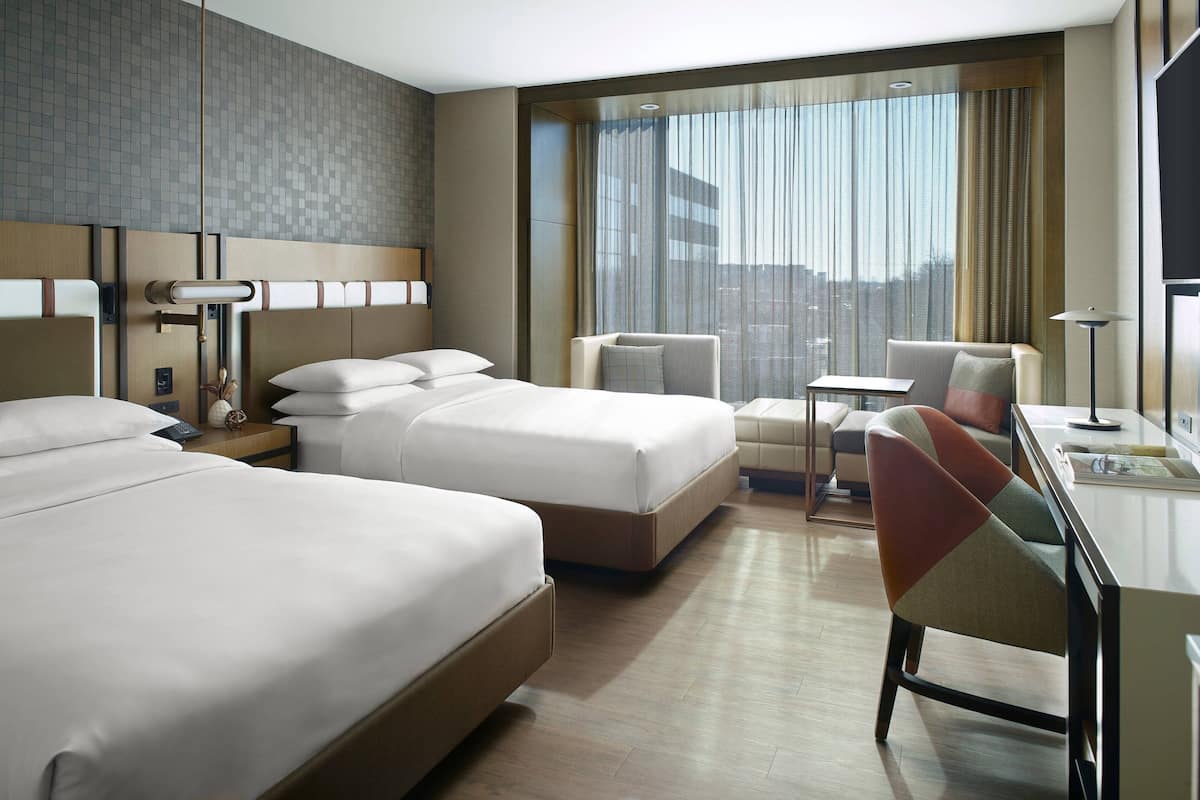 A hotel room with two beds, a desk, a chair, and a view of the city through large windows.