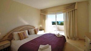 Une chambre d’hôtel avec un grand lit, une table de chevet, une lampe, une vue sur l’extérieur par une fenêtre et un balcon.