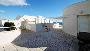 Une terrasse sur le toit, avec un escalier menant à un niveau supérieur, un bâtiment blanc et une vue sur la mer.