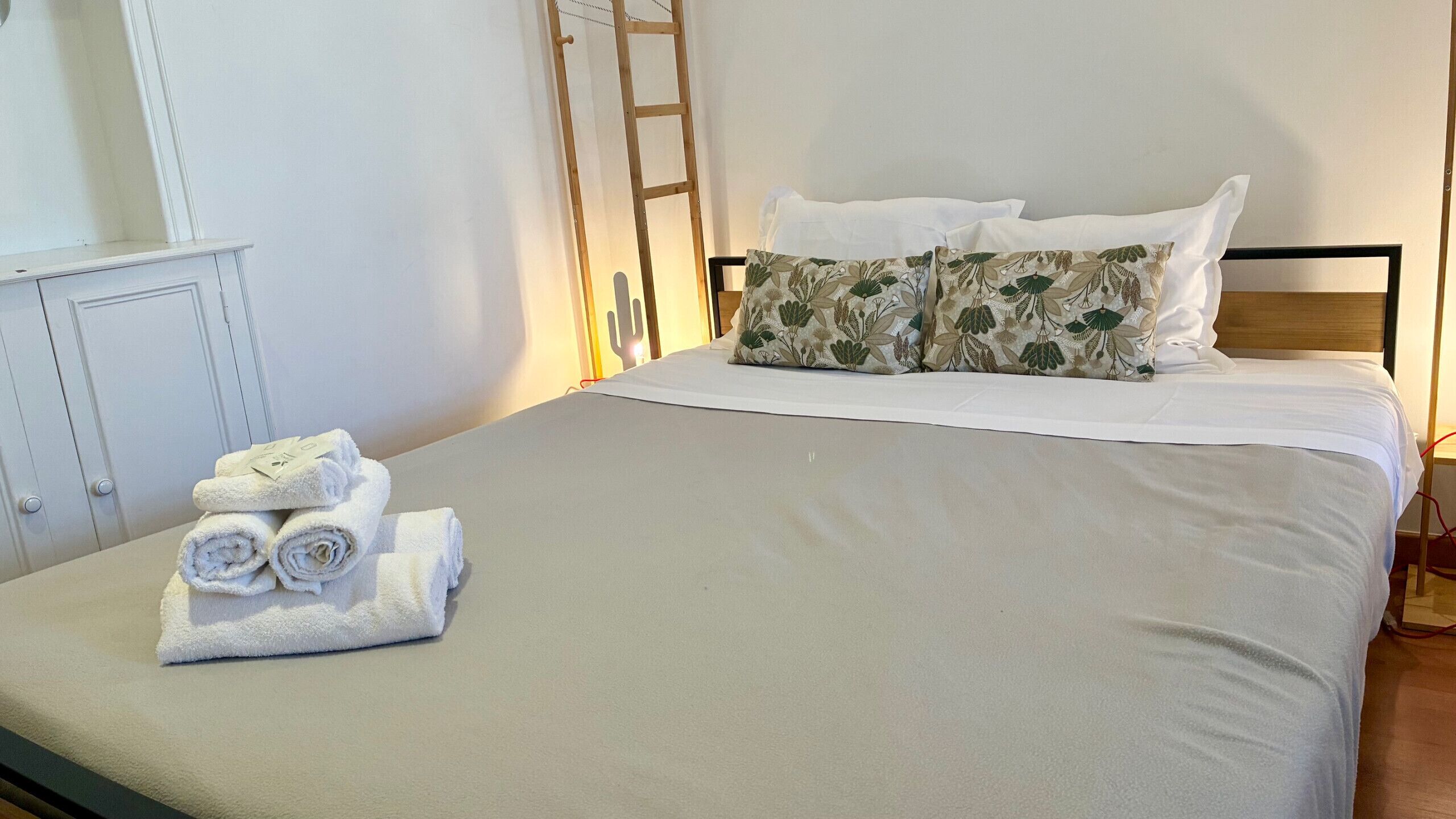 A neatly made bed with white linens and floral-patterned pillows, a stack of folded towels, and a wooden ladder against the wall.