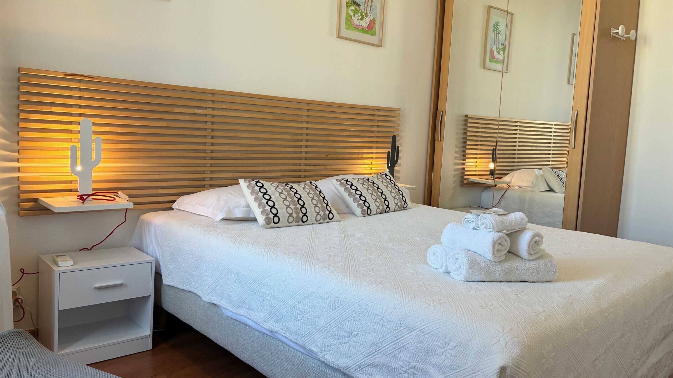 A neatly made bed with white linens, a cactus-shaped night light, and patterned pillows. There is a bedside table with a remote control and a small drawer. A stack of folded towels is on the bed.