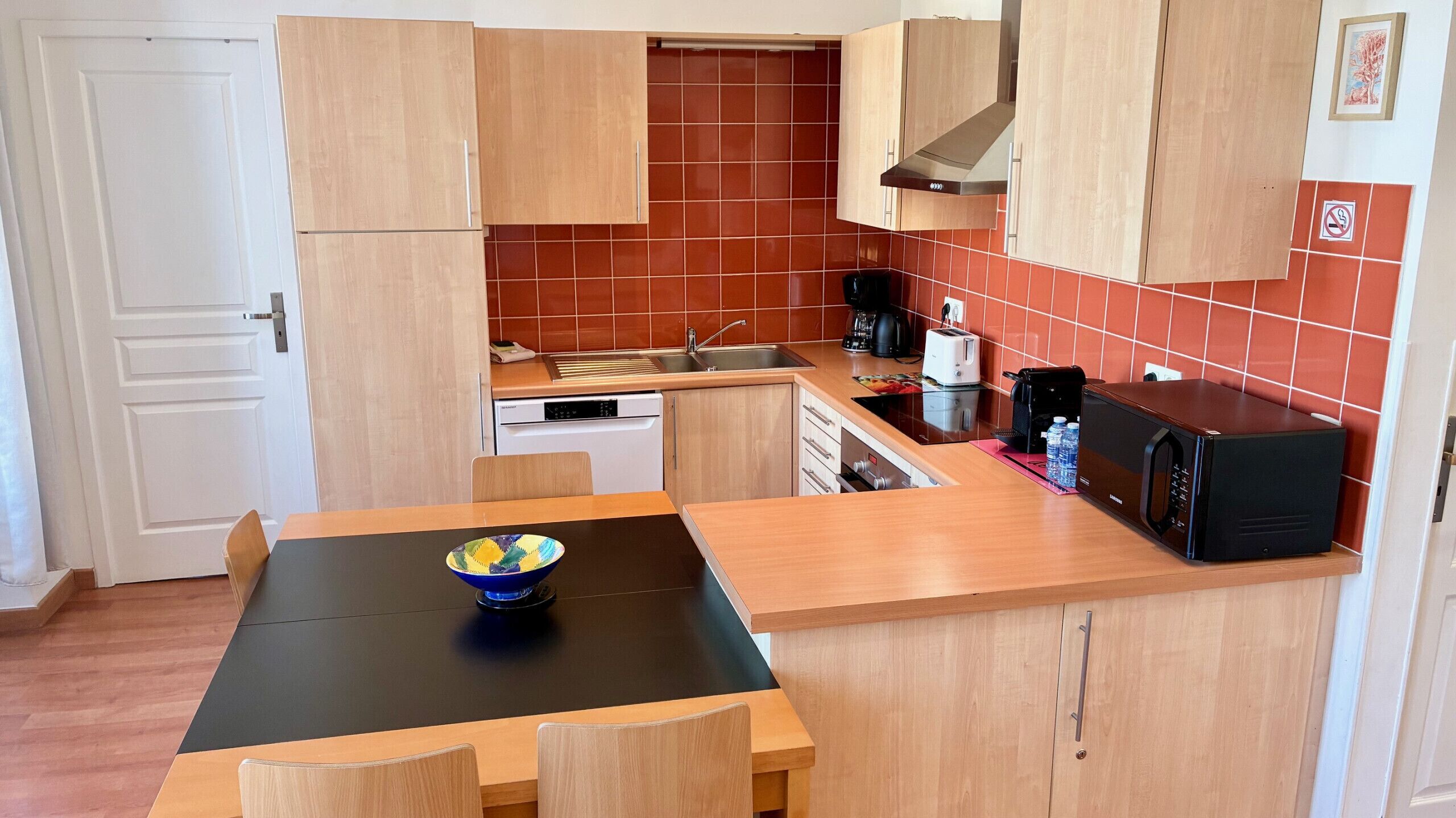 Una cucina compatta con mobili in legno, un piano di lavoro nero, un lavandino, una lavastoviglie, un forno a microonde e un tavolo con sedie.