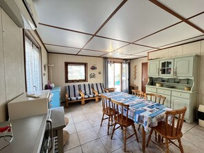 A dining area with a long table covered in a tablecloth, surrounded by wooden chairs. There is a kitchen area with green cabinets and a window.