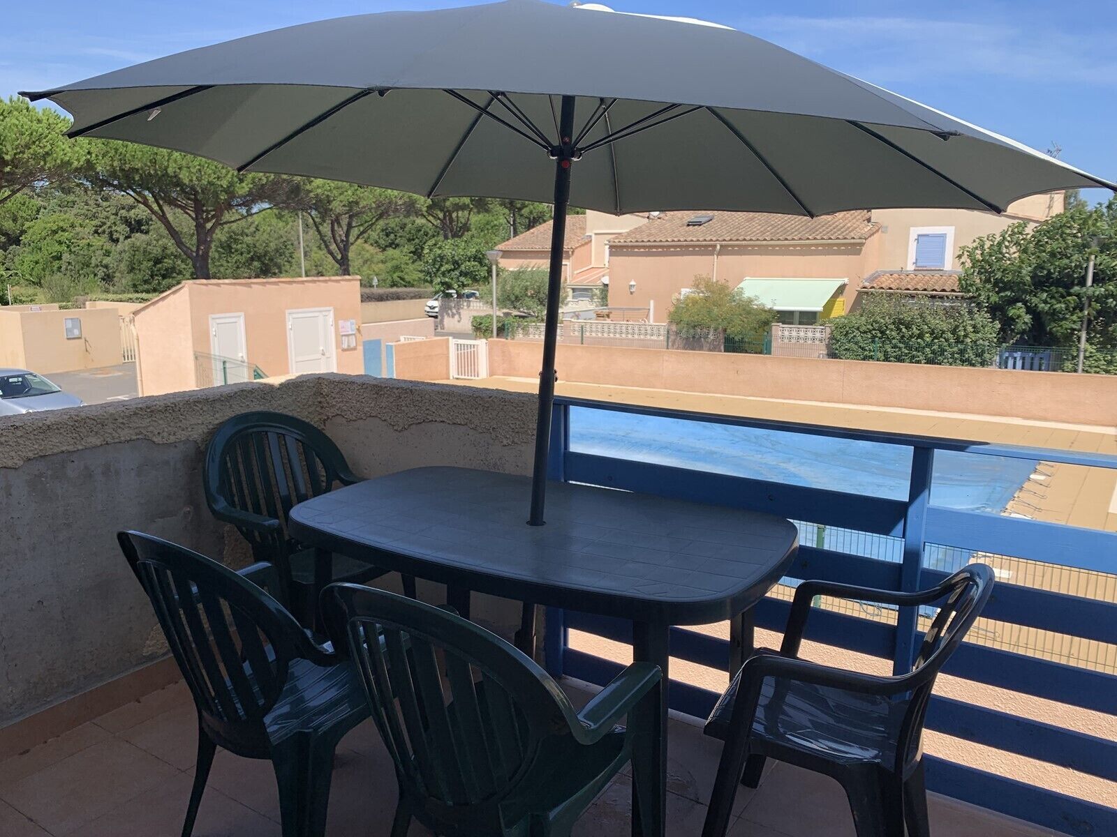 A balcony with a table and chairs under a large umbrella, overlooking a swimming pool and residential buildings.