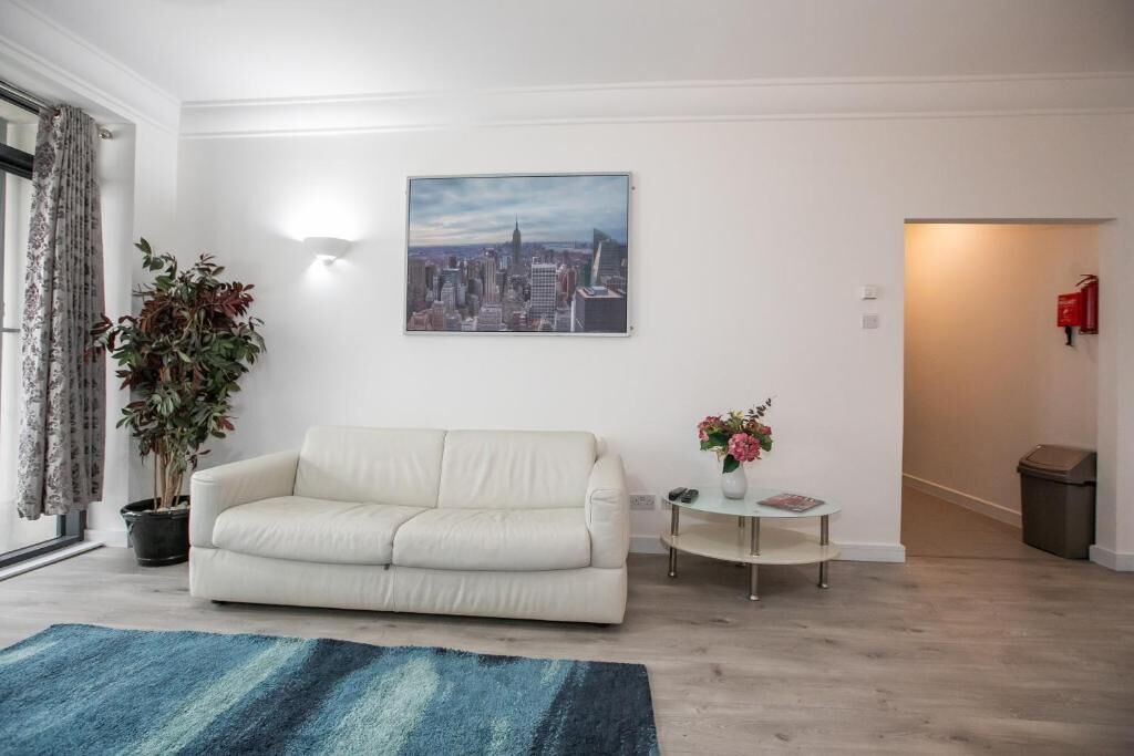 A living room with a white sofa, a blue rug, a cityscape painting, a potted plant, and a small table with a vase of flowers.
