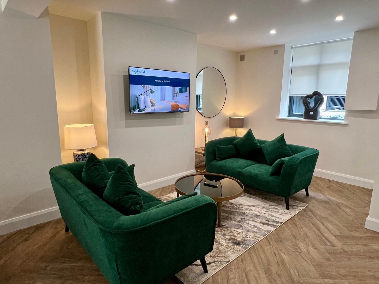 A modern living room with two green armchairs, a coffee table, a flat-screen TV, a mirror, and a window with a view.