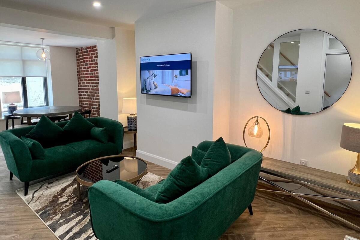 Un salon moderne avec des canapés verts, un téléviseur fixé au mur, un miroir rond et une petite table.