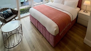 A modern bedroom with a large bed, a chair, a small table, and a view of the city through large windows. - IFSC Townhouse Apartments (Dublin)