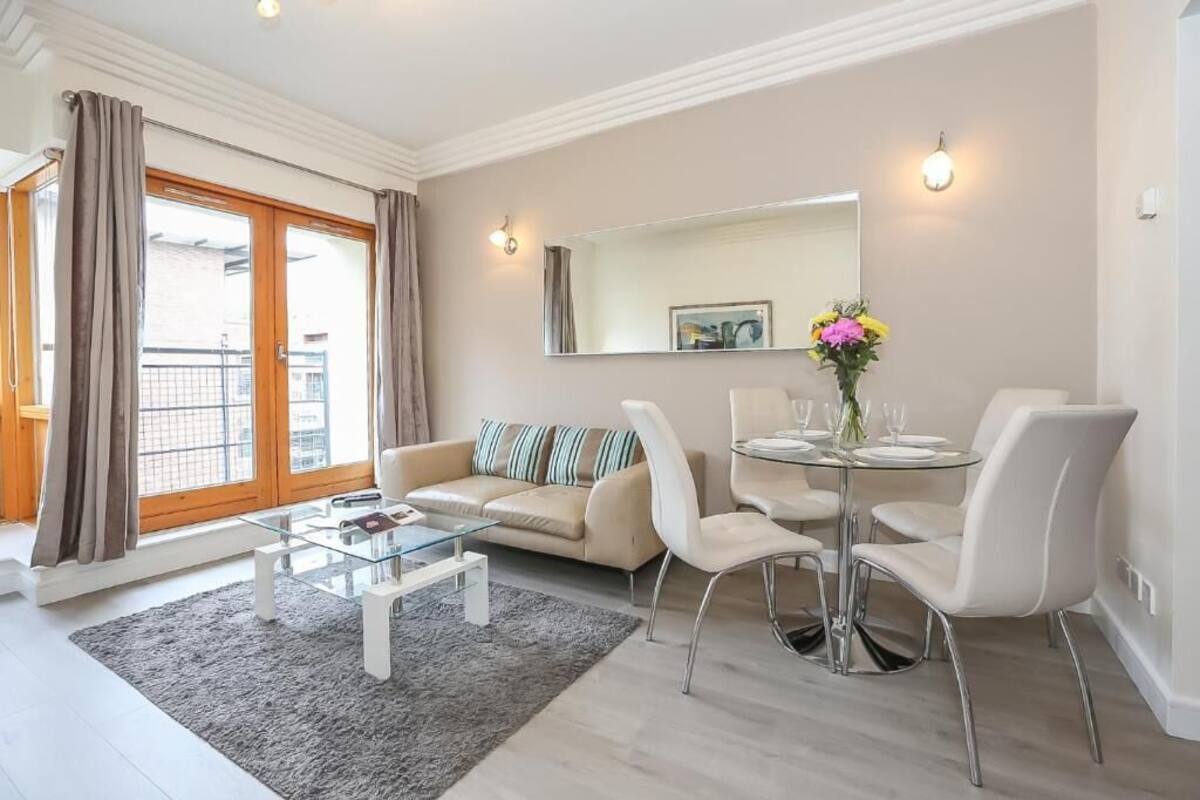 Un salon moderne avec une table basse en verre, un canapé beige agrémenté de coussins à rayures, une table à manger avec des chaises blanches et une grande fenêtre avec des rideaux.
