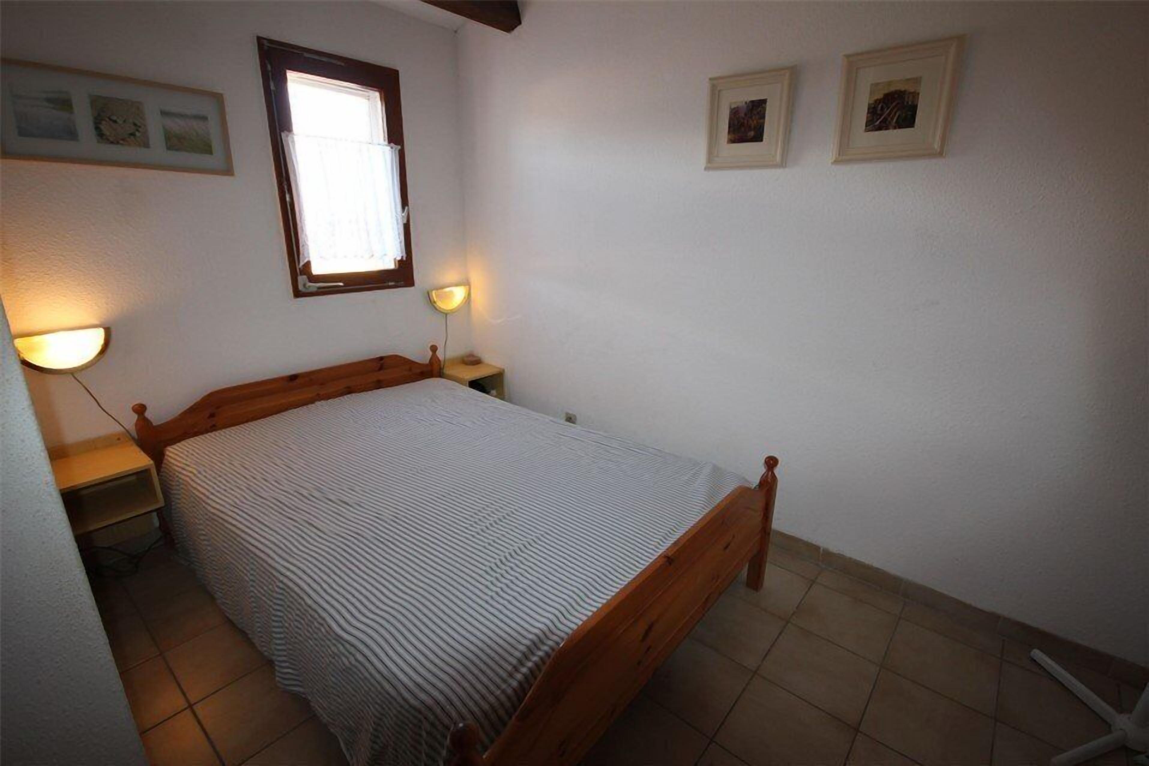 A bedroom with a wooden bed, two bedside lamps, a window, and two framed pictures on the wall.