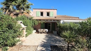 Uma casa com telhado de telhas, um alpendre com mesa e cadeiras e uma pequena área de jardim com palmeiras.