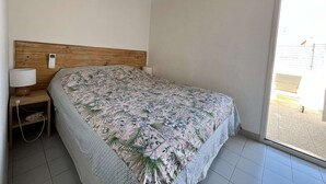 Un dormitori amb un capçal de fusta, un llit amb estore floral, una taula de nit amb una làmpada i una finestra amb vista exterior.