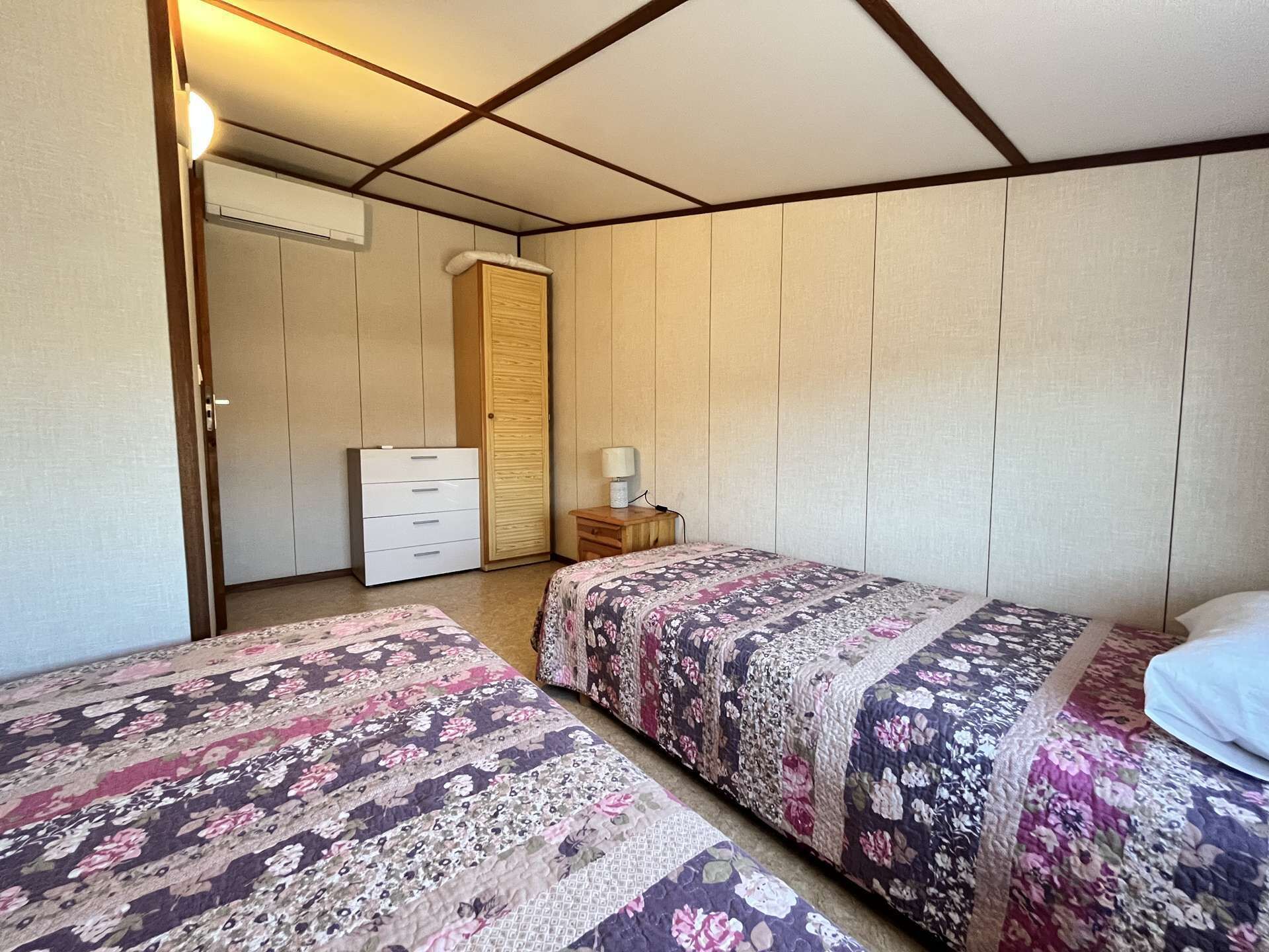 A bedroom with two beds, a wardrobe, a bedside table with a lamp, and a ceiling fan.