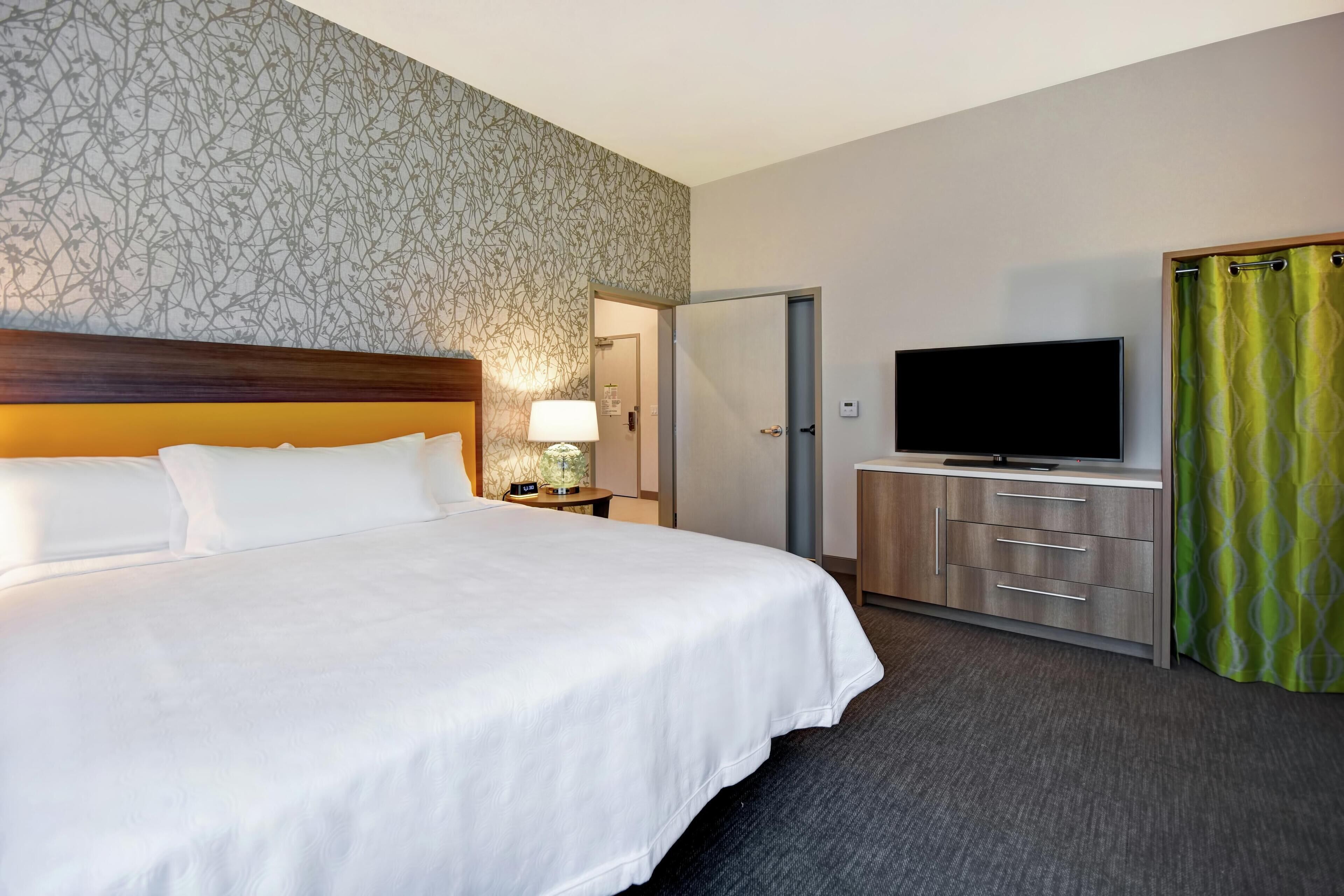 Een hotelkamer met een groot bed, een televisie op een houten dressoir, een nachtkastje met een lamp, en een behang met patroon.
