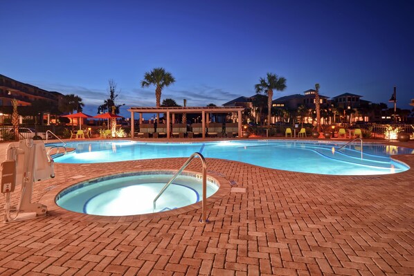 Zona de piscina con jacuzzi, rodeada de un patio de ladrillo, palmeras y un área de estar cubierta con sombrillas.