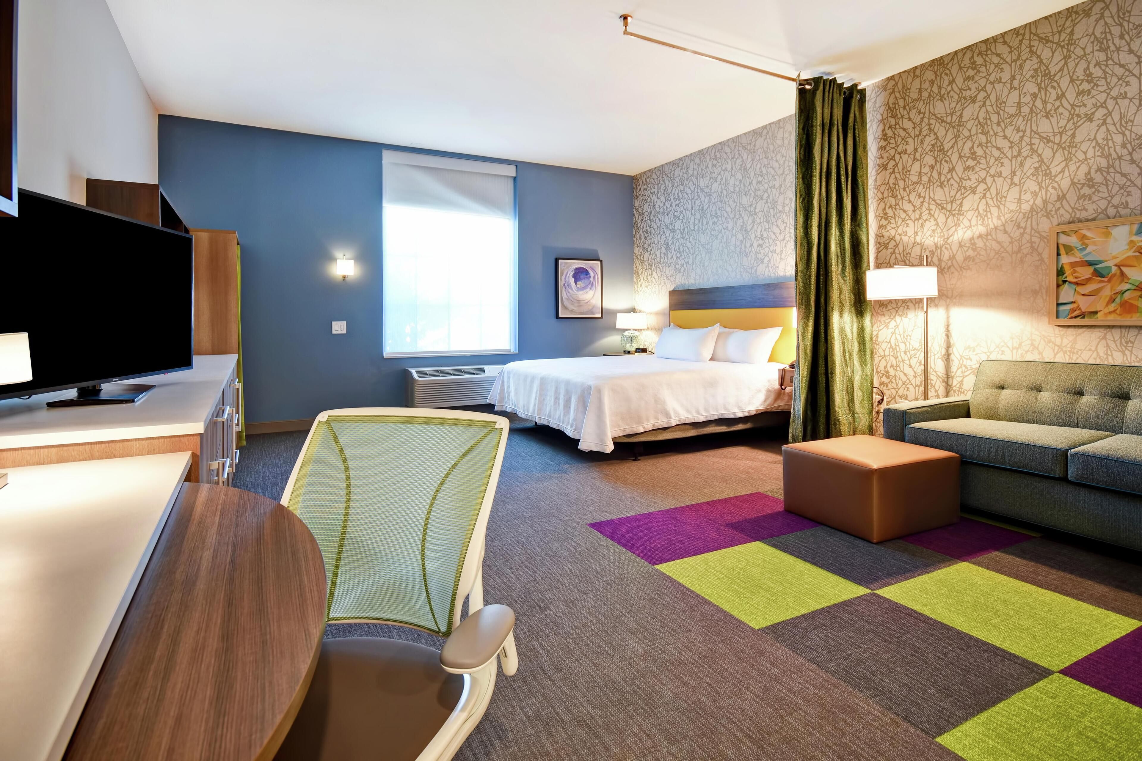 Une chambre d’hôtel avec un lit, un canapé, un bureau avec une télévision, une chaise et une moquette colorée.