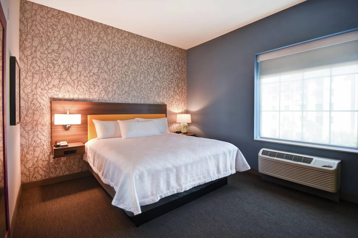 A hotel room with a bed, nightstands, wall-mounted lamps, a window, and an air conditioning unit.