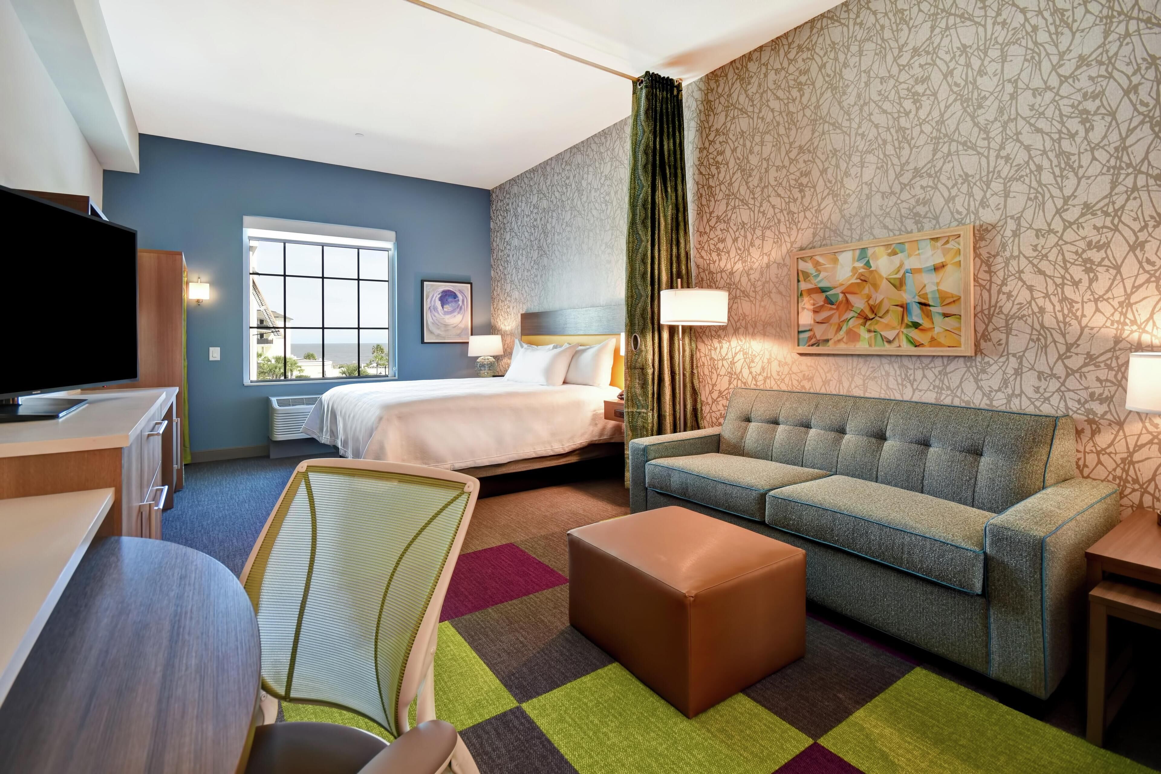 A hotel room with a bed, a sofa, a TV, a desk, and a colorful rug.