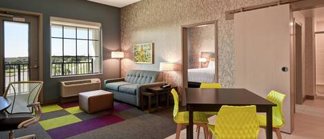 A modern hotel room with a sofa, a dining table with chairs, a small table with a lamp, and a window with a view of greenery.