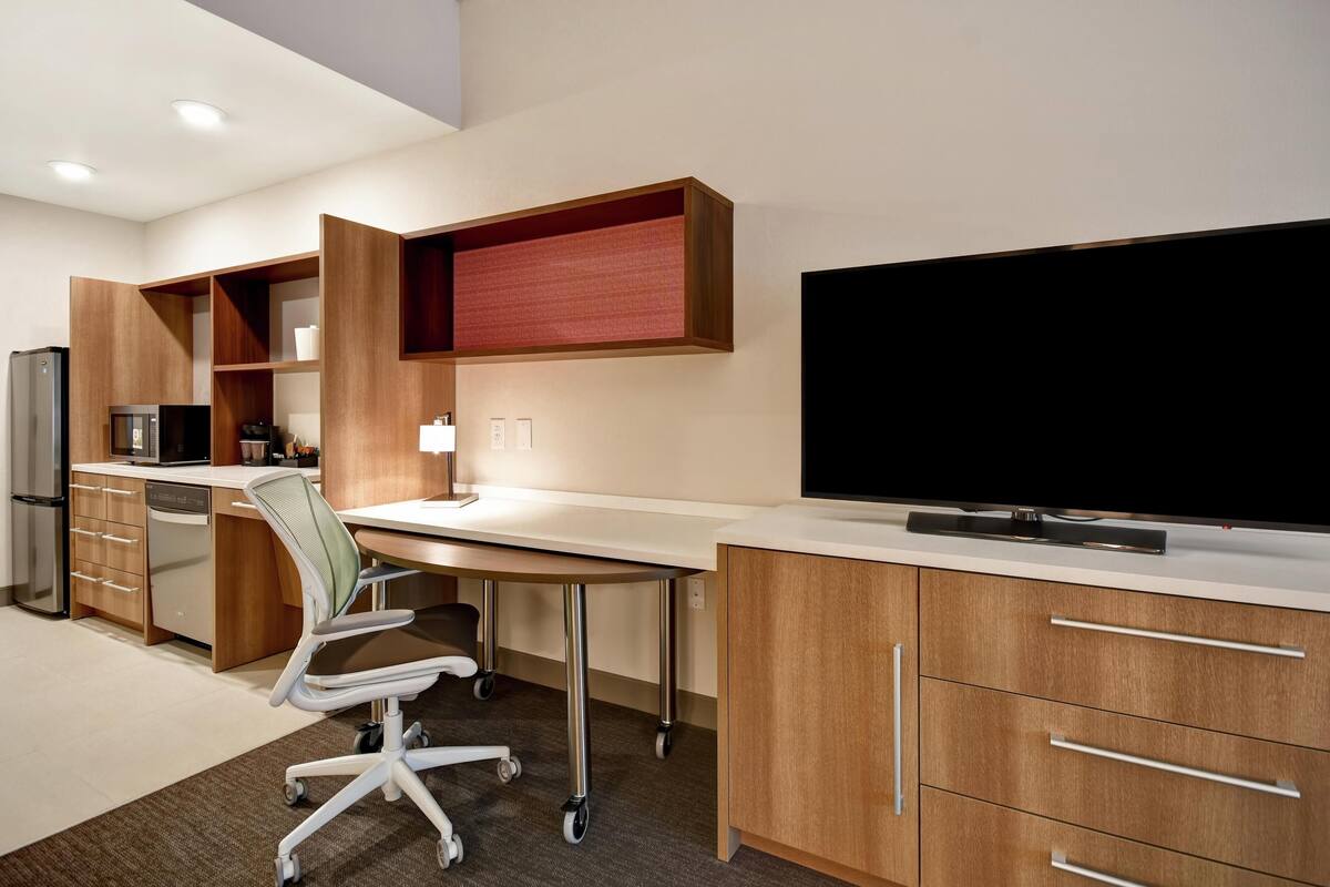A modern kitchen with wooden cabinets, a large flat-screen TV, a kitchen island with a chair, and a refrigerator.