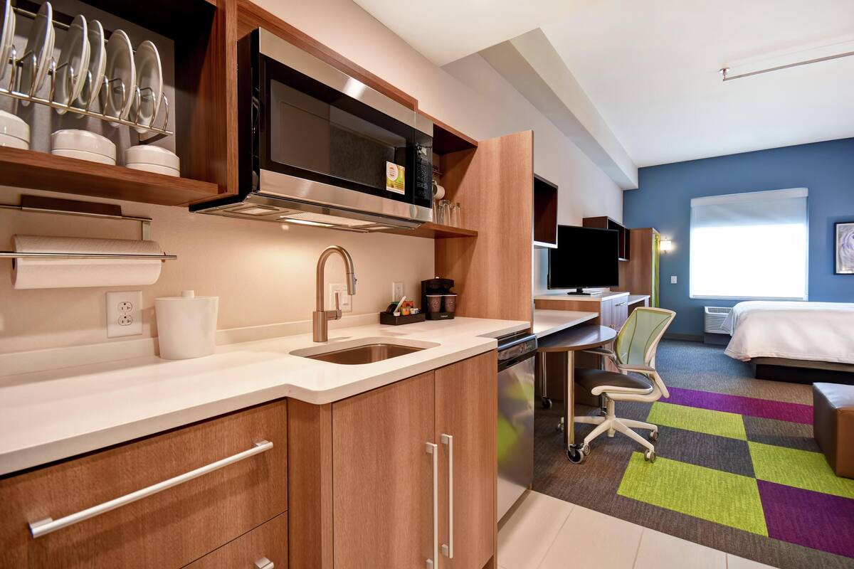 A modern kitchen with a microwave, sink, and wooden cabinets. There is a dining area with a chair and a bed in the background.
