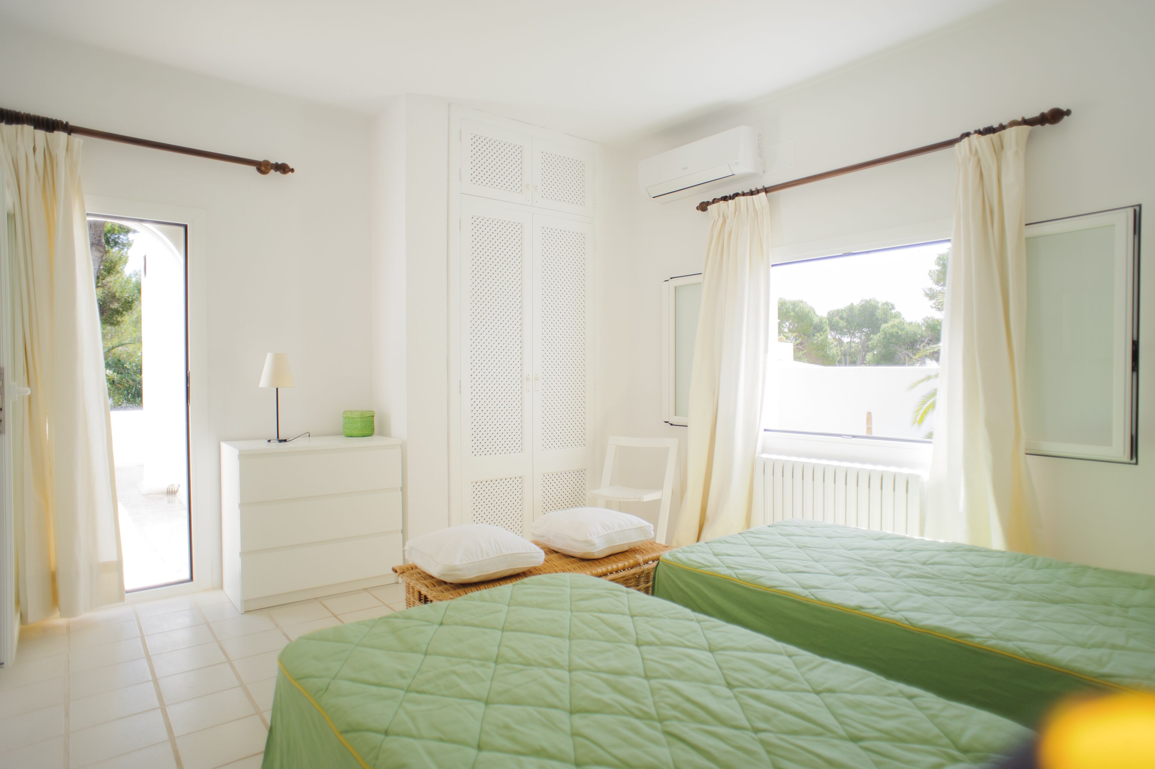 Een slaapkamer met twee bedden, een raam met gordijnen, een witte kledingkast en een klein tafeltje met een lamp.