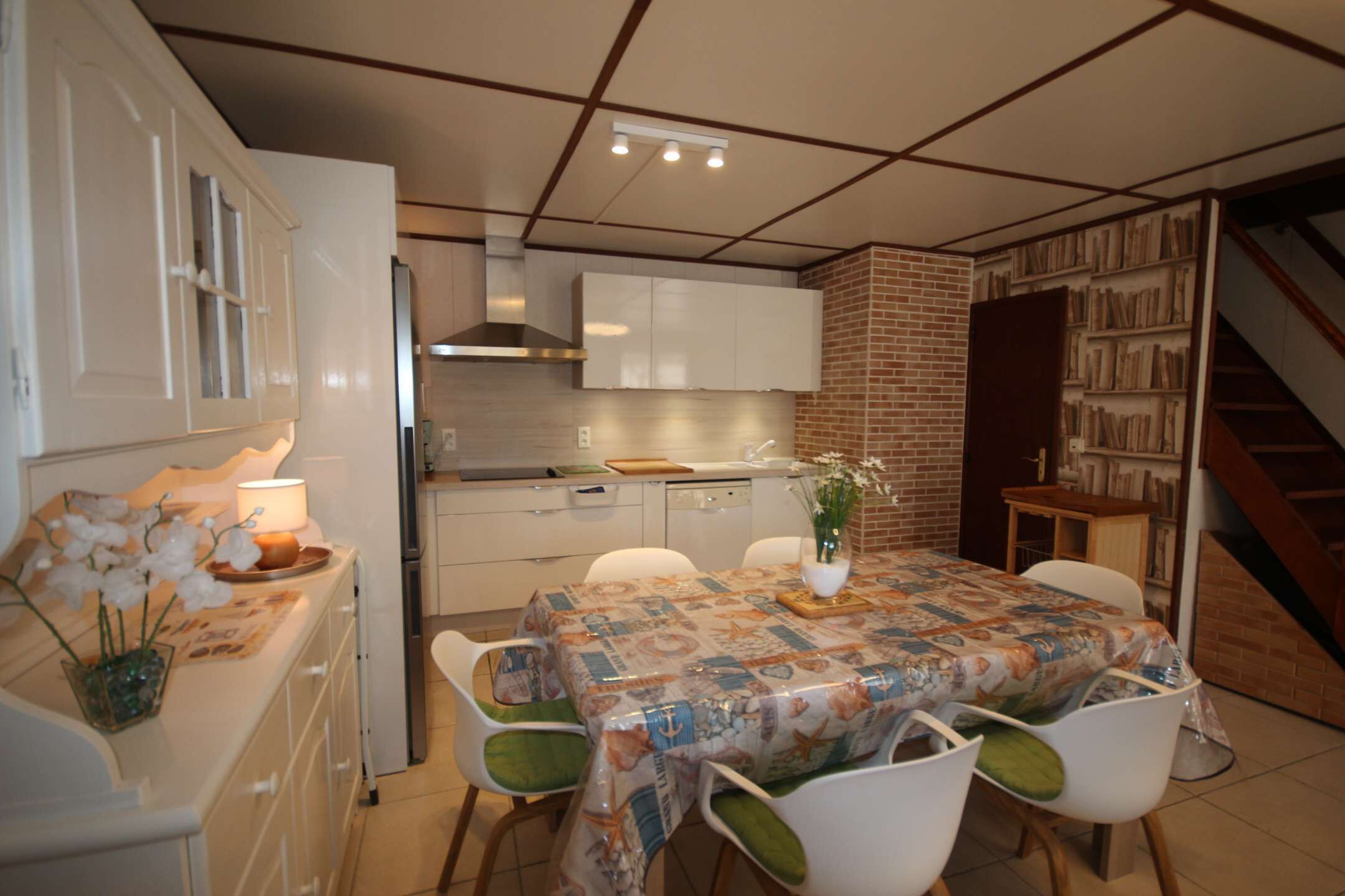 Une cuisine compacte avec des meubles blancs, une table à manger recouverte d’une nappe à motifs et un escalier menant à un espace rempli de livres.
