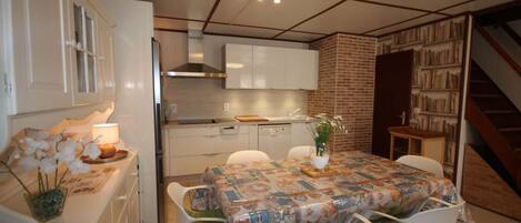 Une cuisine compacte avec des meubles blancs, une table à manger recouverte d’une nappe à motifs et un escalier menant à un espace rempli de livres.