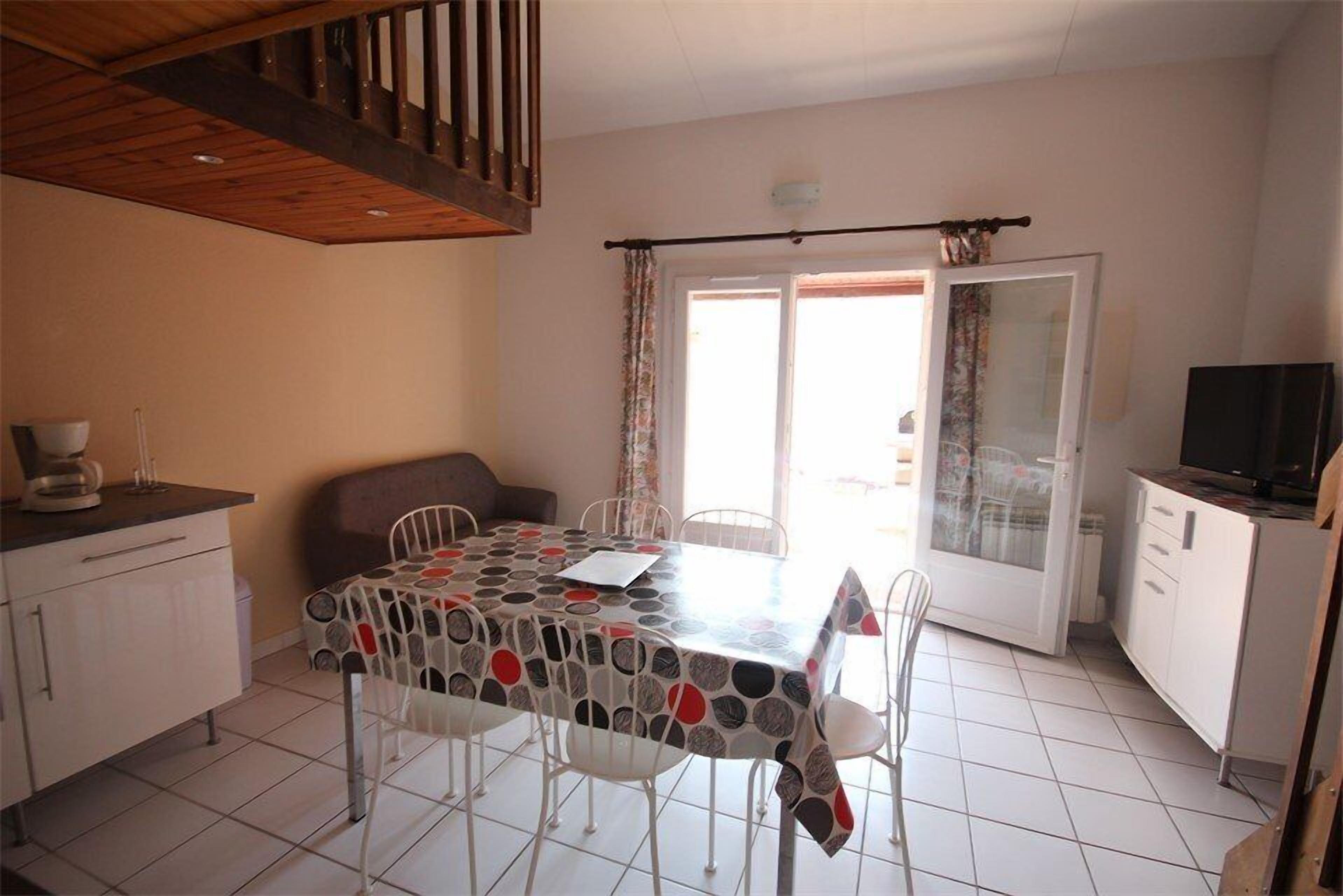 A dining area with a table covered by a patterned tablecloth, surrounded by white chairs. There is a kitchen area with cabinets and a television on a stand.