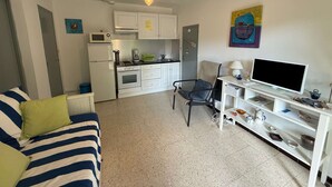 En kompakt opholdsstue med et tekøkken, en sofa, en lænestol, et fjernsyn og en reol med forskellige genstande.