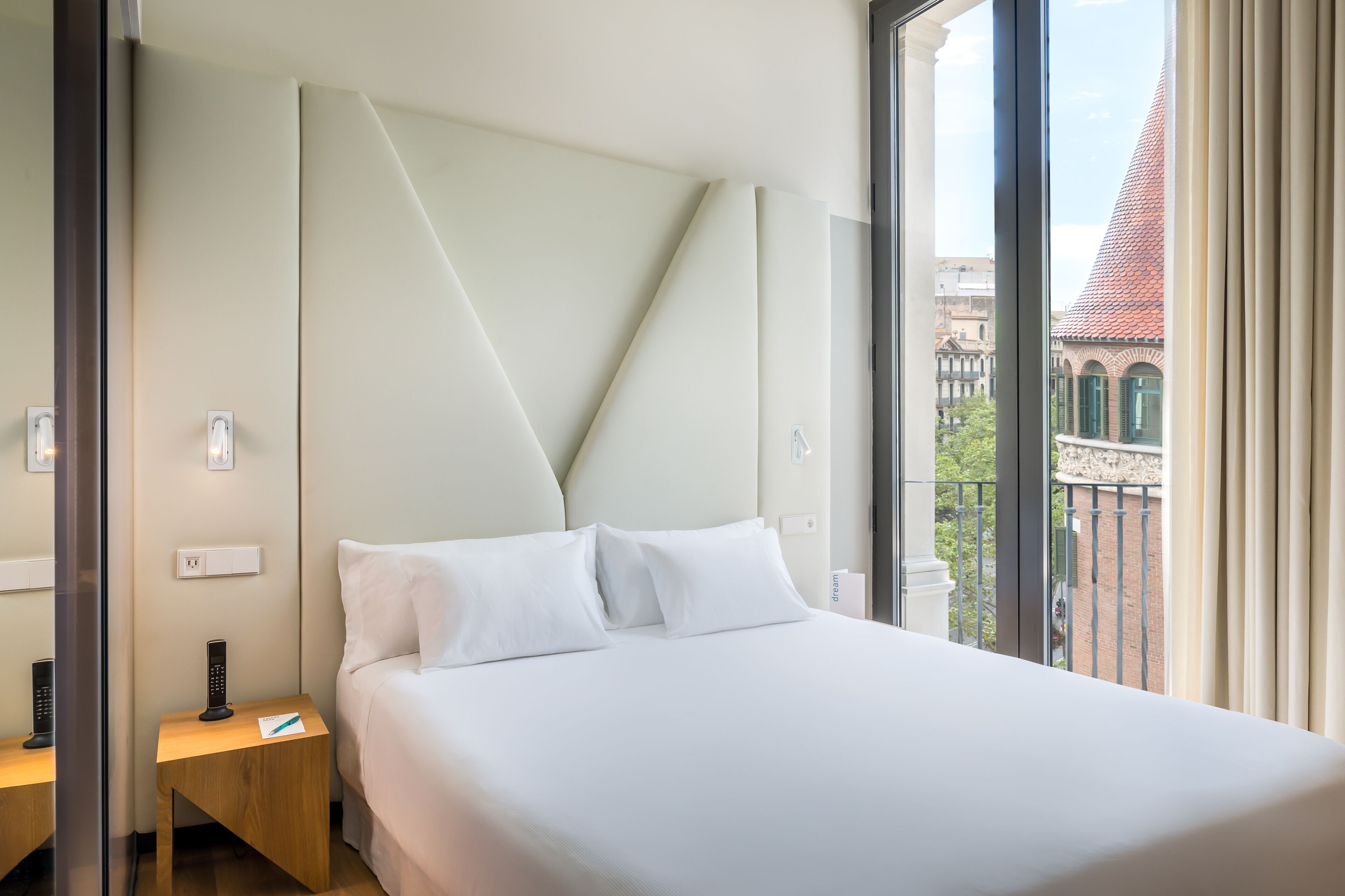 A hotel room with a large bed, a bedside table with a remote control, a wall-mounted light, and a view of a building with a red roof.