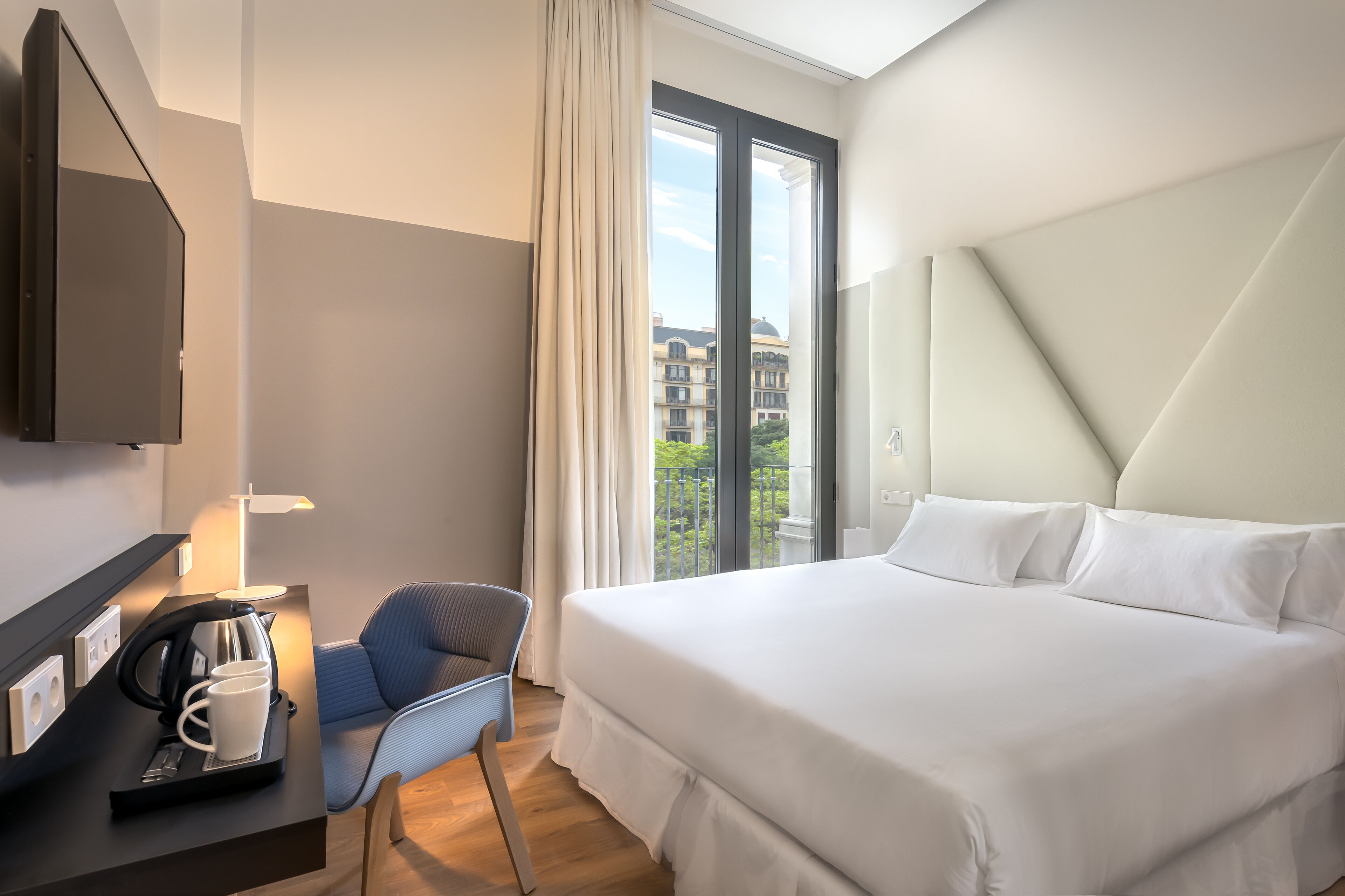 A hotel room with a large bed, a desk with a coffee maker, a chair, and a view of buildings through the window.