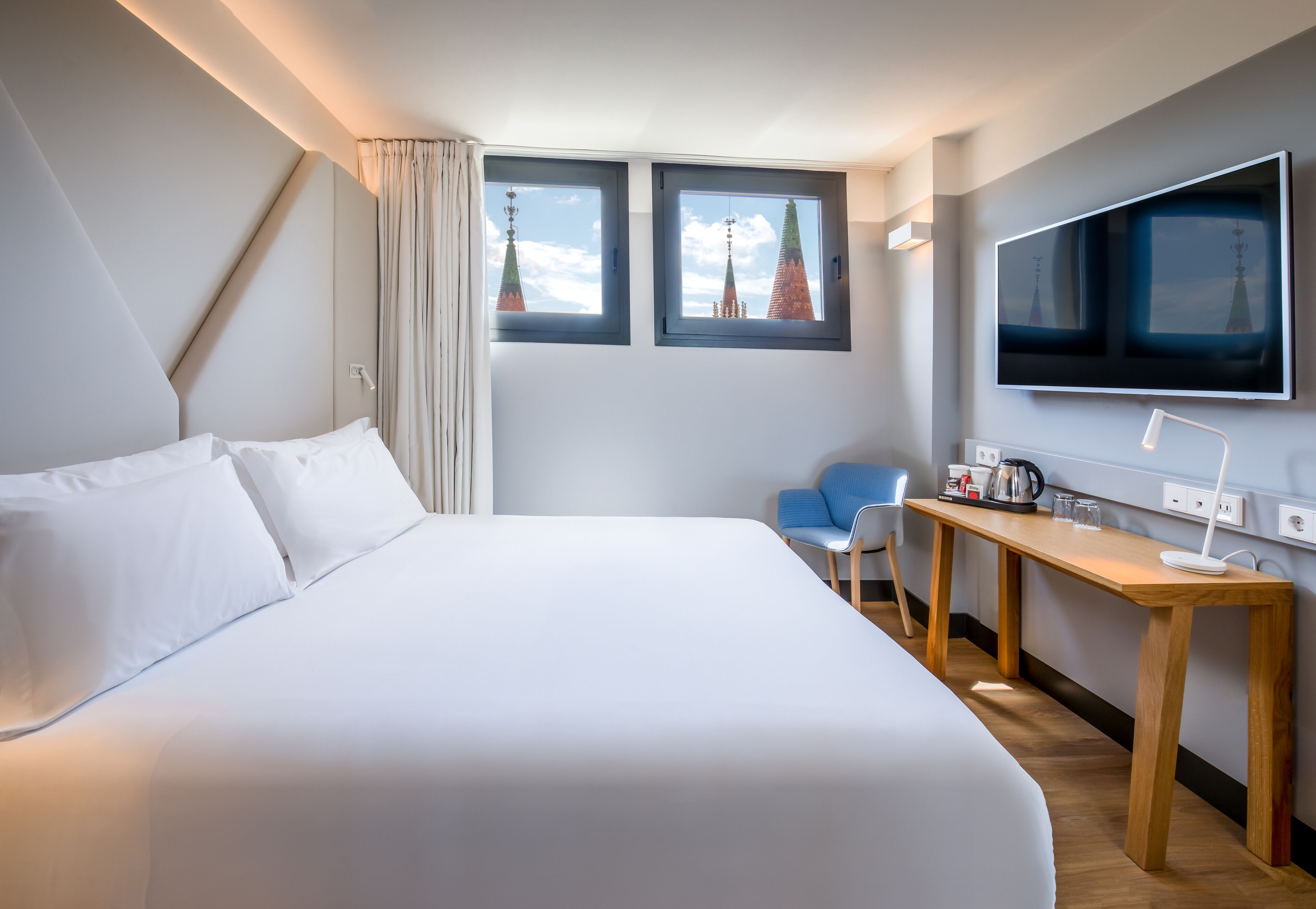A hotel room with a large bed, a desk, a chair, a TV, and two windows with a view of buildings.