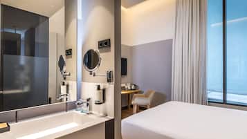 Een moderne hotelkamer met een grote spiegel, een witte wastafel, een bed, een bureau en uitzicht op de stad door het raam.