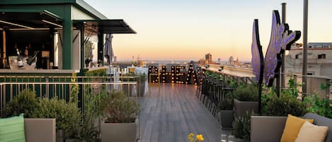 Una terrazza sul tetto con vista sullo skyline cittadino al tramonto, con posti a sedere all'aperto, un'area bar e illuminazione decorativa.