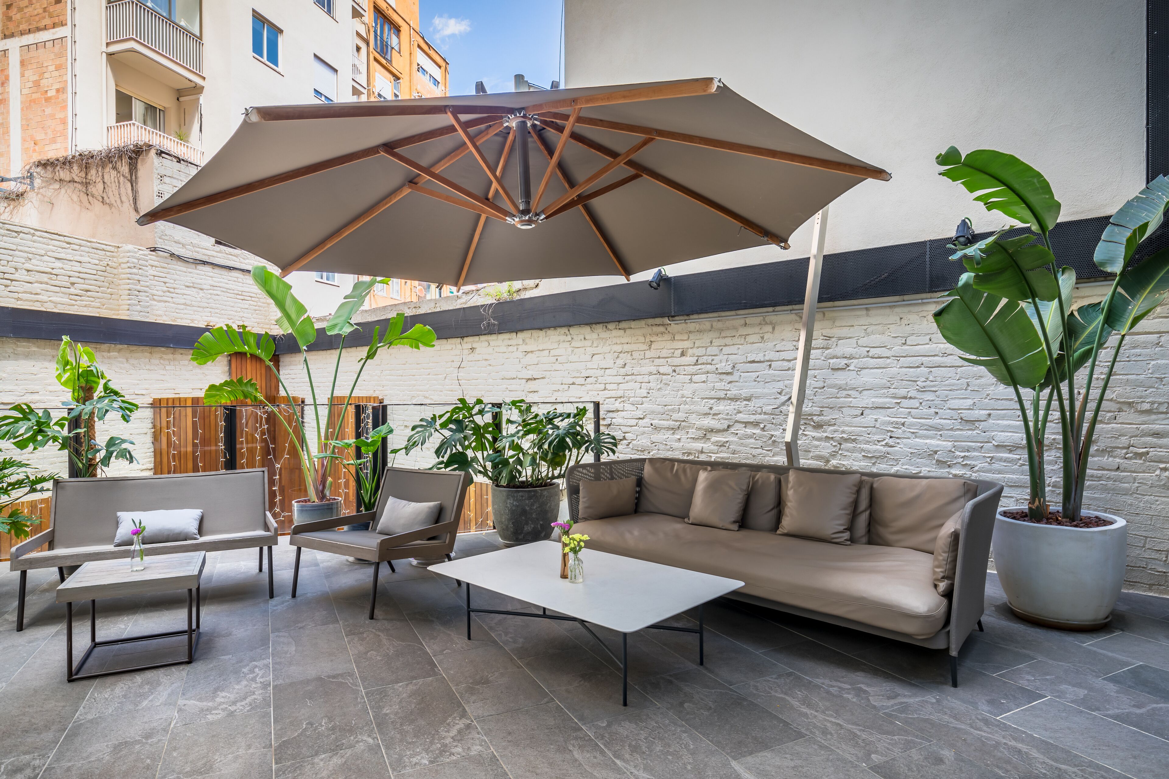 An outdoor patio with a sofa, two chairs, a coffee table, and a large umbrella. There are potted plants and a brick wall in the background.