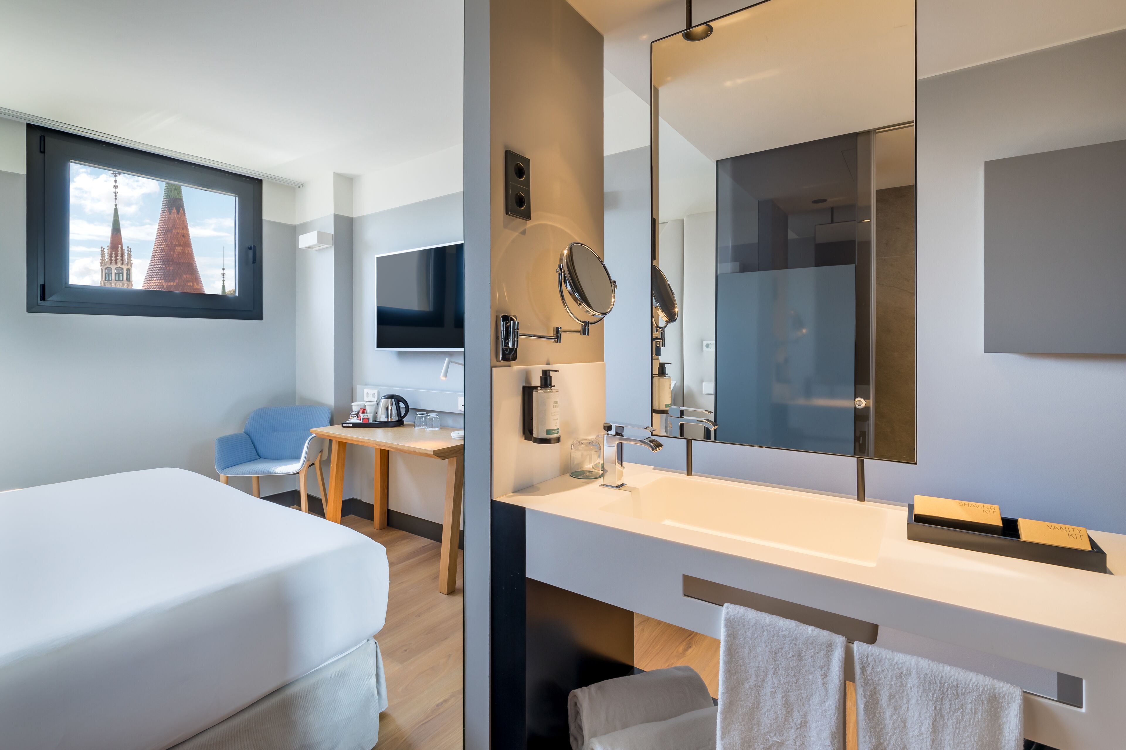 Ein modernes Hotelzimmer mit einem Bett, einem Schreibtisch, einem großen Spiegel und Blick auf ein Gebäude durch ein Fenster.