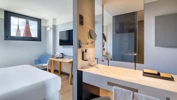 A modern hotel room with a bed, a desk, a large mirror, and a view of a building through a window.