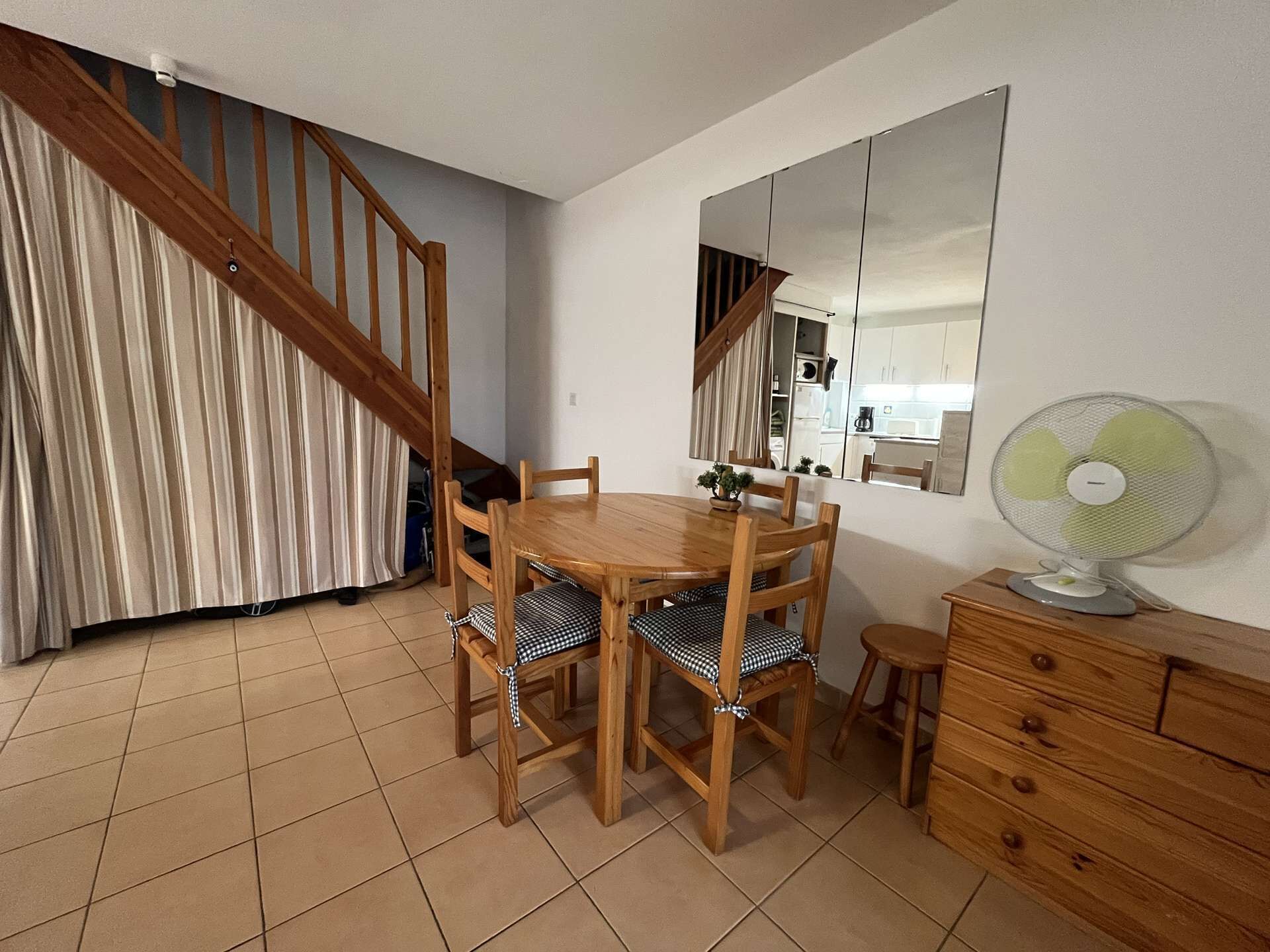 Uma área de refeições com uma mesa e cadeiras de madeira, um espelho, um ventilador e uma escada.