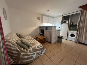 A compact living space with a sofa, a small table, a kitchen area including a microwave and washing machine, and a trash bin.
